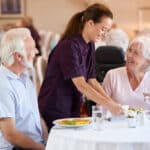 Senior Couple Being Served With Meal By Carer In Dining Room Of Retirement Home
